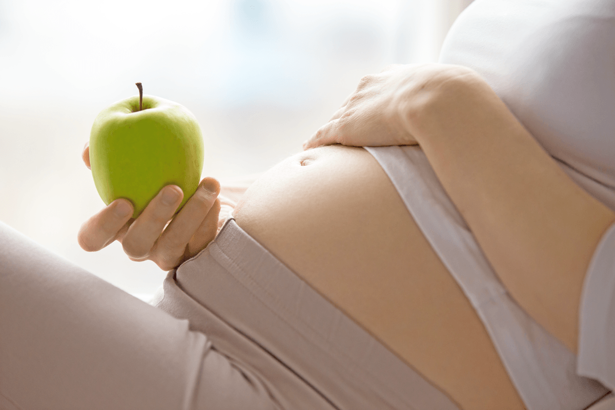 Ishrana u trudnoći - trudnica leži na krevetu, njen trudnički stomak je jasno vidljiv,a u desnoj ruci drži zelenu jabuku.