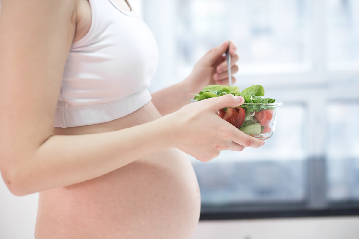 Ishrana u trudnoći - trudnica nosi običnu belu majicu koja otkiva njen trudnički stomak. U rukama, u staklenoj činiji, joj je salata od povrća sa zelenom salatom, paradajzom.